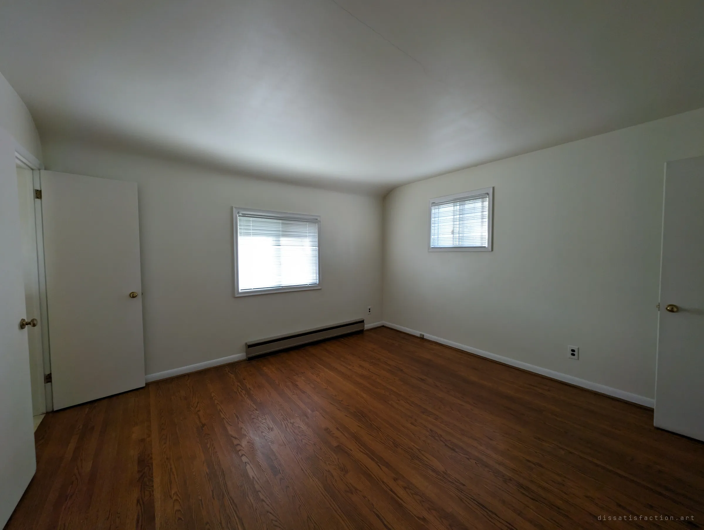 An empty bedroom with bare hardwood floors, a baseboard heater and blinds drawn over two windows. Clean and untouched, as if waiting for the next owner.