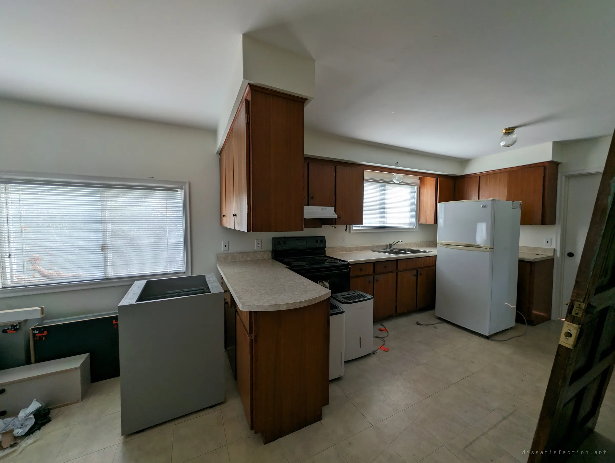 A dated kitchen with wood cabinets, a white fridge and a half-installed grey cabinet box in the middle of the floor. Renovation work seems to have stopped partway.
