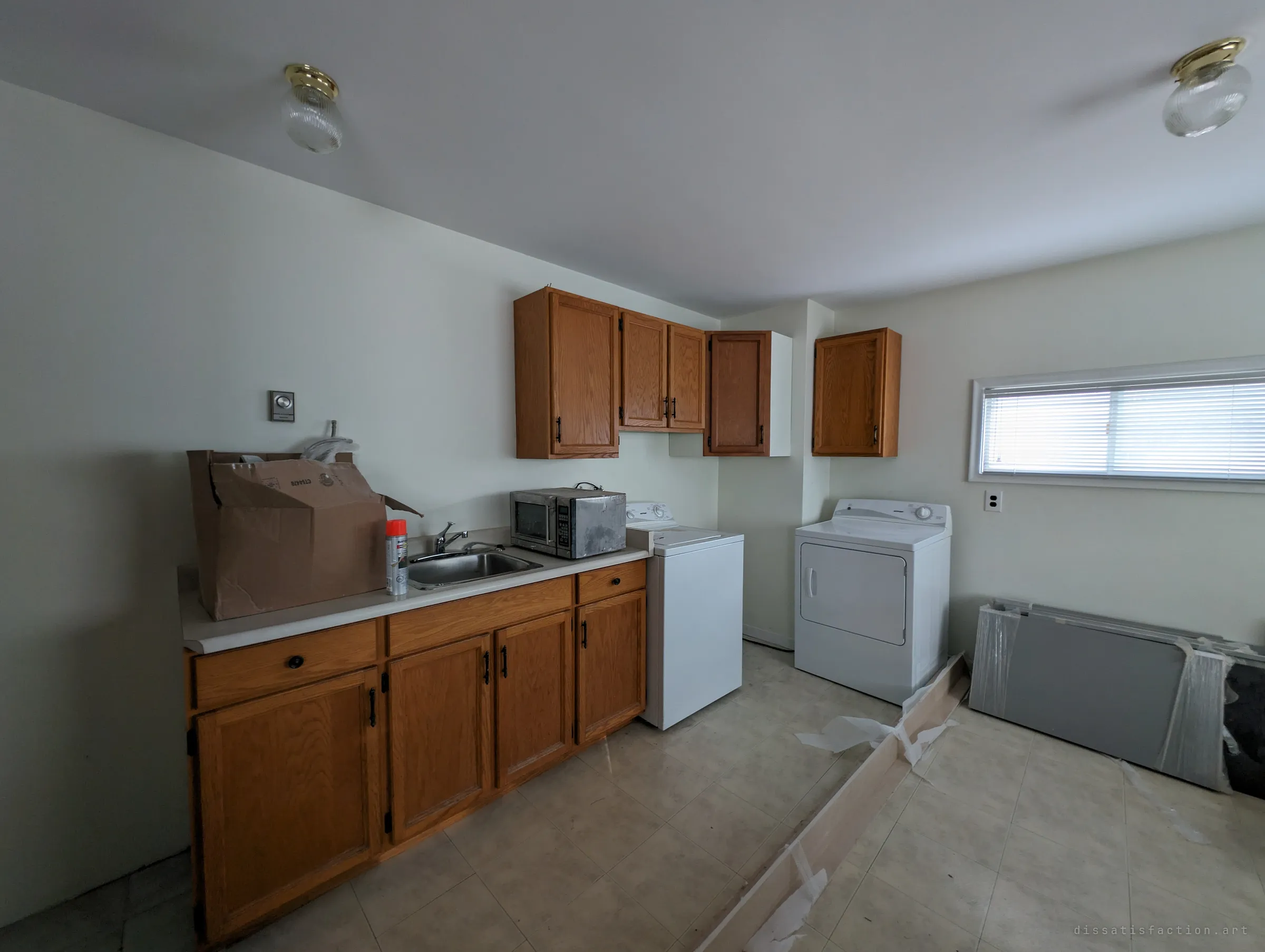 A laundry room with oak cabinets, a sink, a dusty microwave, a washer and dryer, and wrapped panels leaning on the wall. Boxes and a spray can suggest abandoned renovation work.