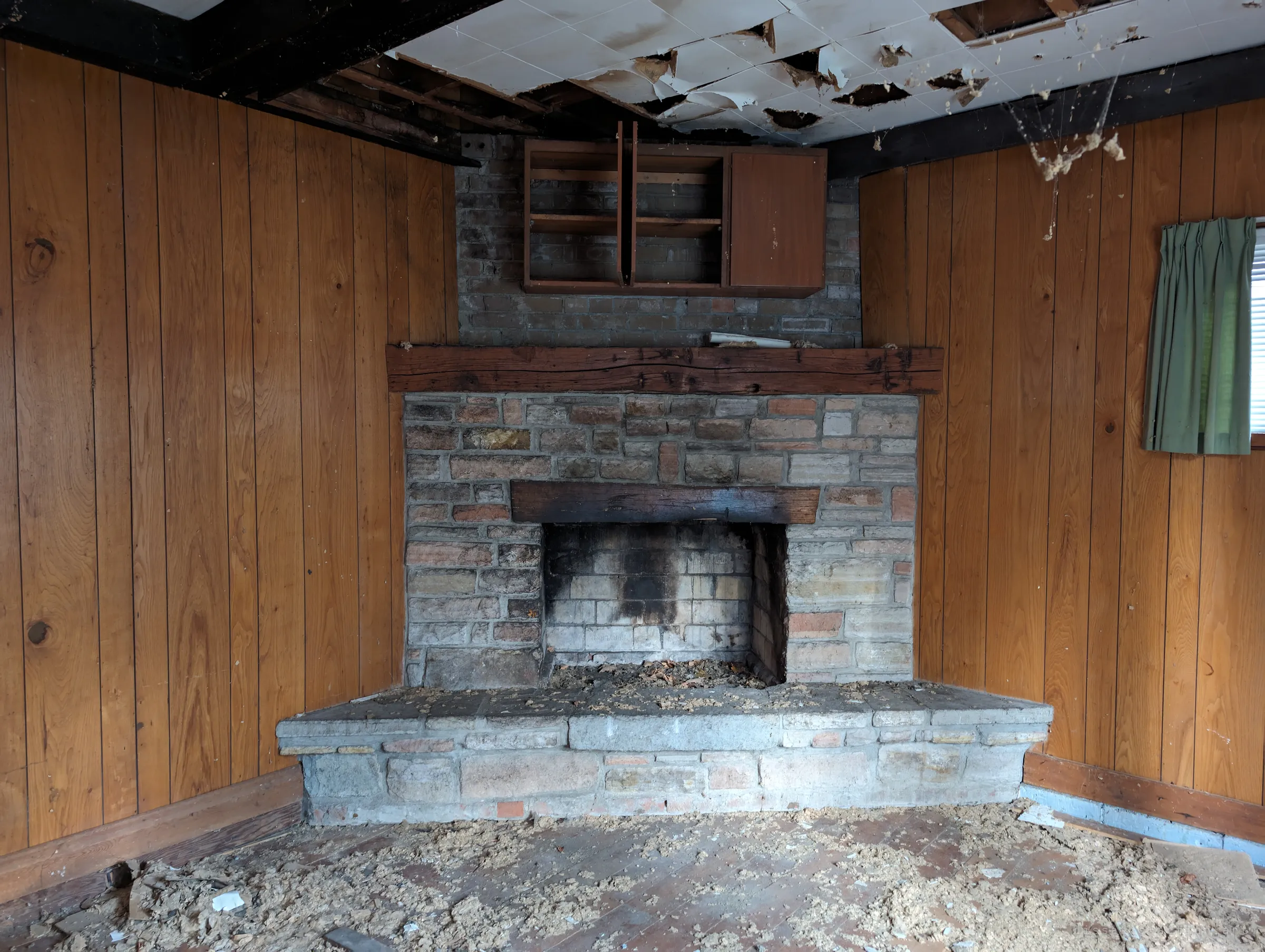 A wood-panelled den with a stone corner fireplace, its ceiling tiles peeling and insulation scattered across the floor. The rougher, older side of the house.