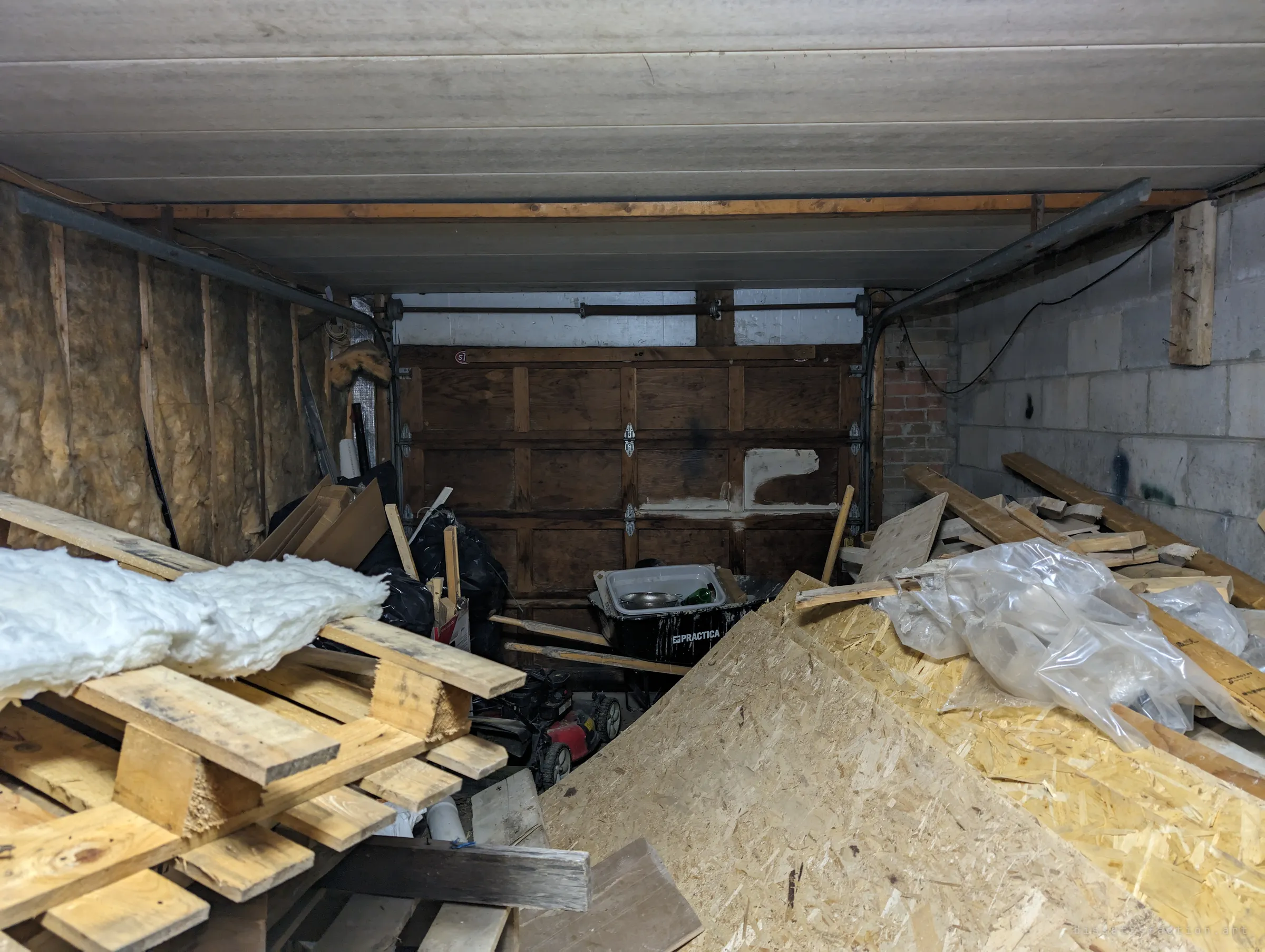 A garage packed with pallets, particle board, insulation and a wheelbarrow in front of an old wooden garage door. Leftover building materials have turned it into a junk pile.