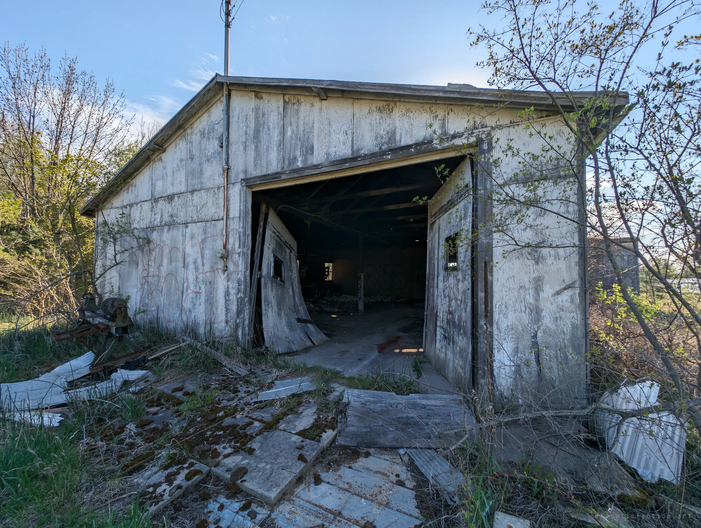 A weathered white garage stands open on a dark, graffiti-marked interior, its sagging doors hanging off their tracks over broken paving slabs. Rusted farm machinery lies to the left.