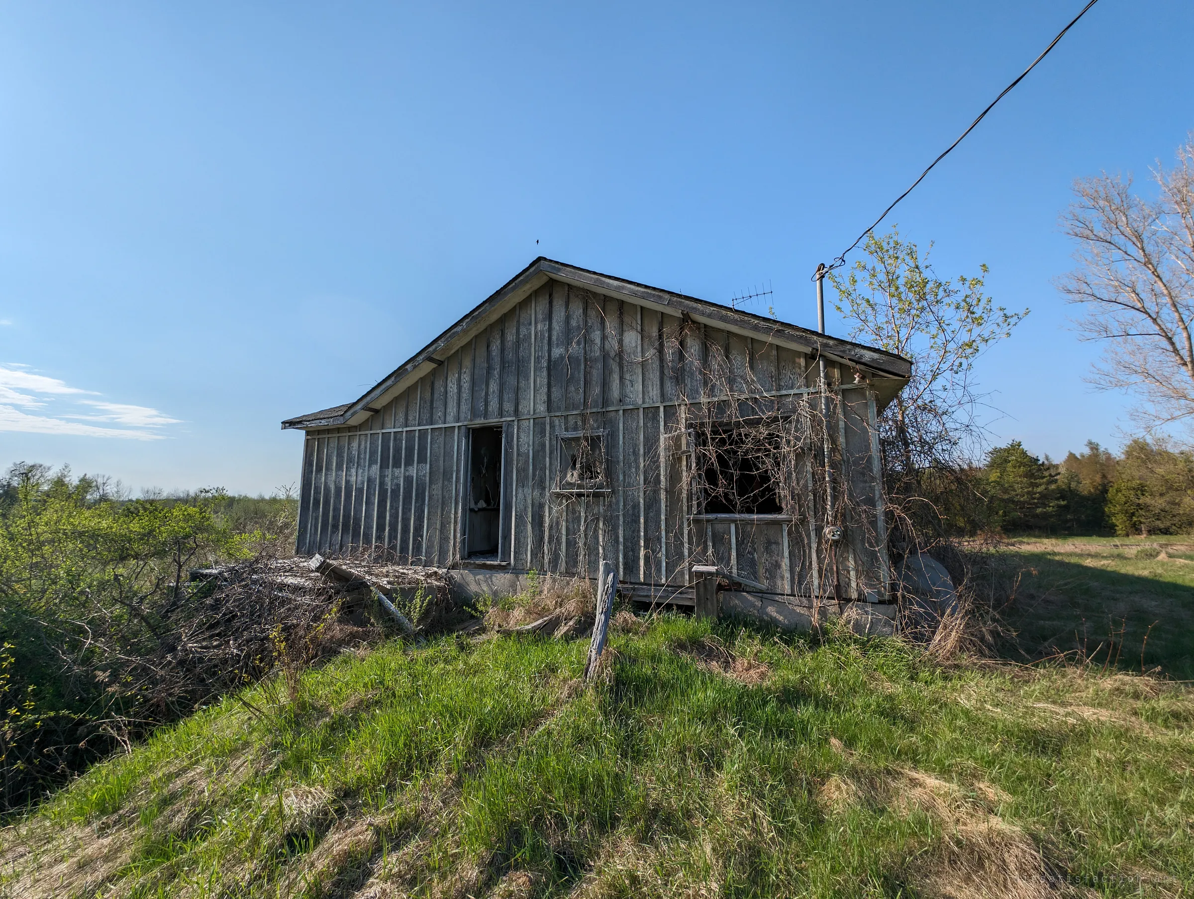 A small grey plank outbuilding perches on a grassy rise under a clear sky, a power line still running to its roof. Open farmland and tree lines stretch behind it.