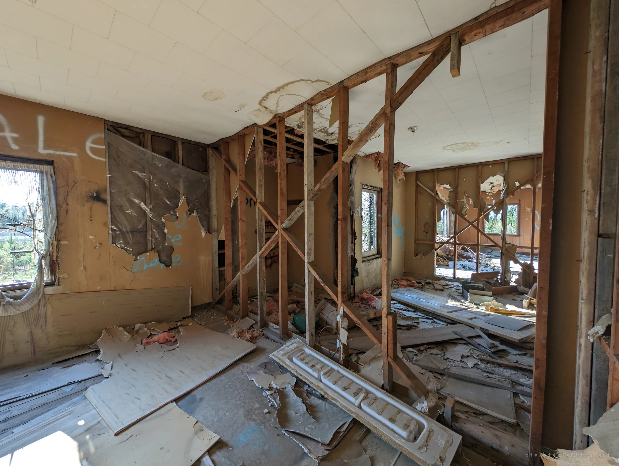A gutted room with walls stripped back to the studs, torn plastic sheeting and graffiti on panelled walls, and debris strewn across the floor. Light floods in through bare window frames.