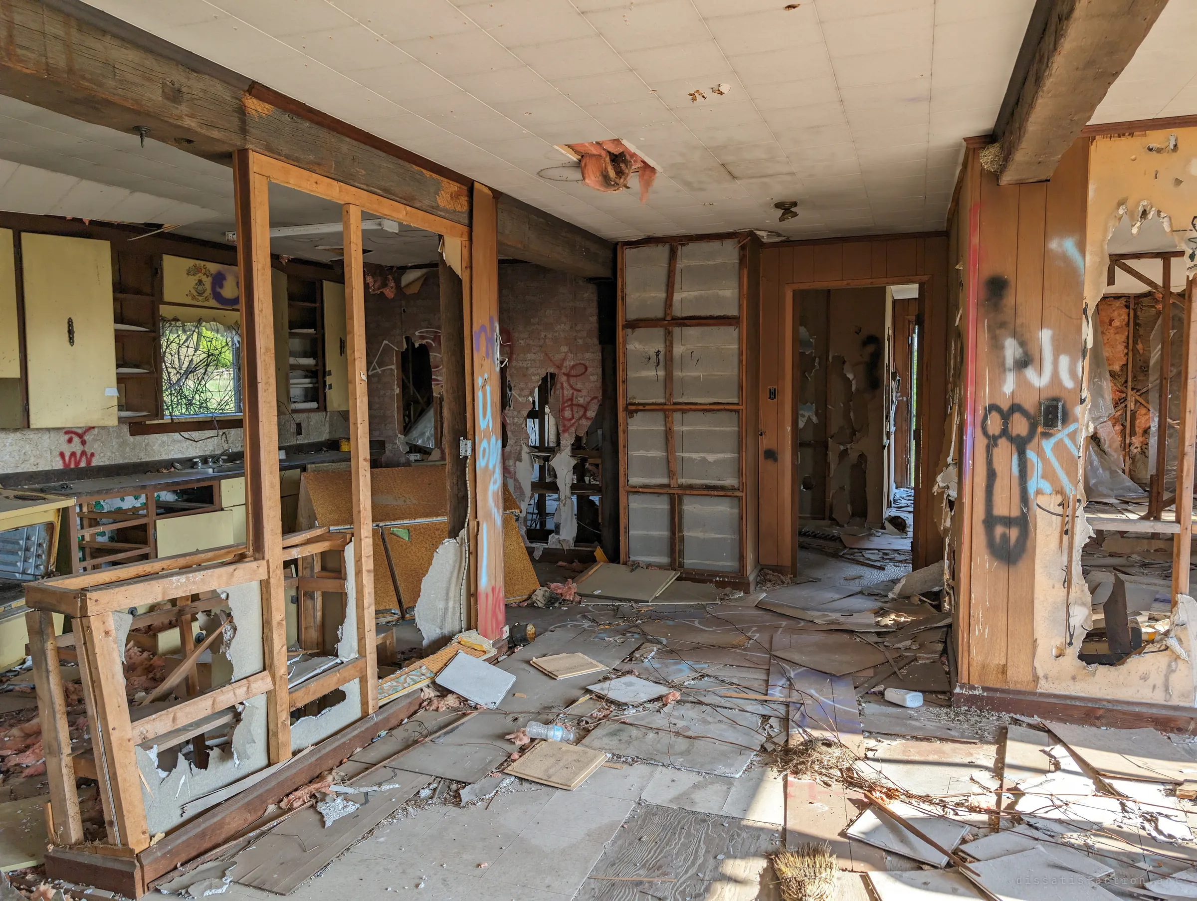 An open-plan living area and kitchen torn apart, with exposed framing, graffiti-covered pillars and fallen ceiling tiles scattered over the floor. Yellow cabinets still hang on the far wall.