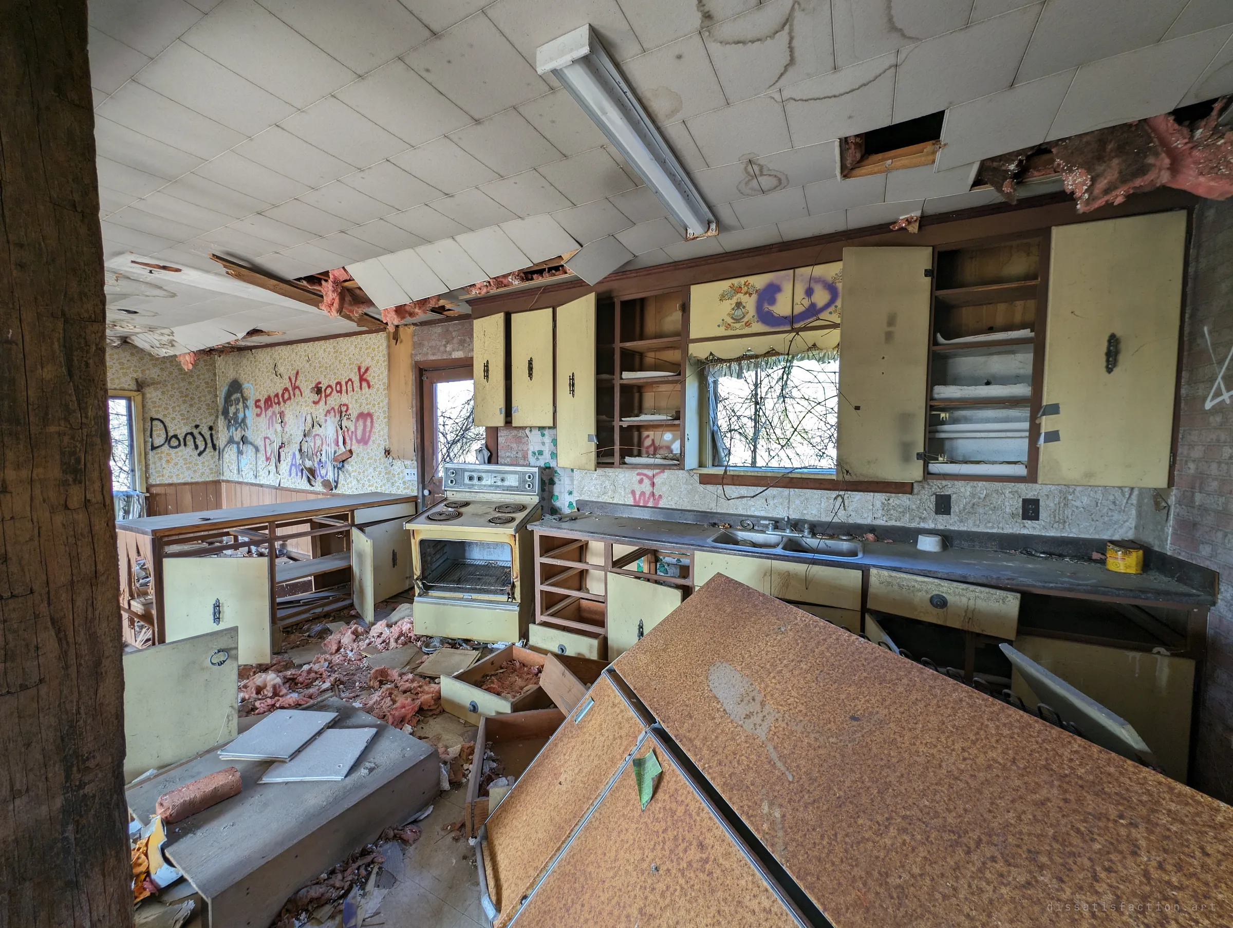 A vintage yellow kitchen with an old electric range, open cabinets and a toppled counter, the ceiling tiles sagging overhead and graffiti on the patterned wallpaper. A frozen slice of mid-century domestic life.