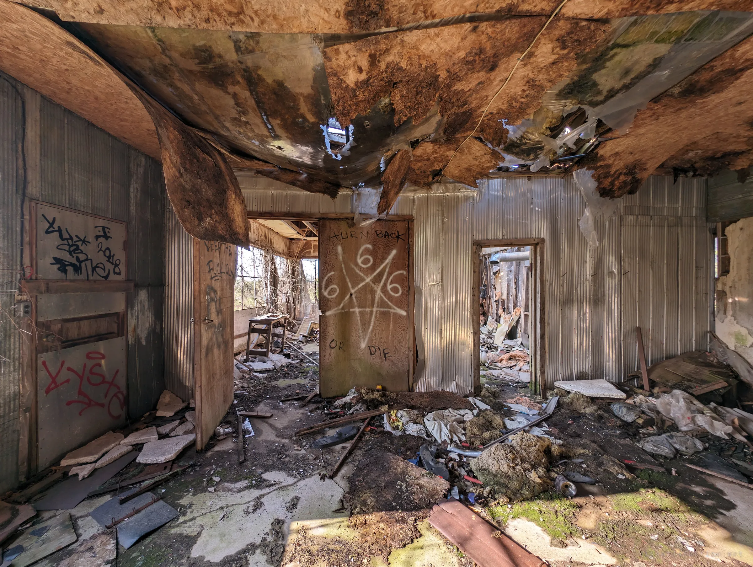 A room with metal-clad walls under a rotting, sagging ceiling, a door spray-painted with a pentagram and a warning to turn back. Debris and moss cover the floor.