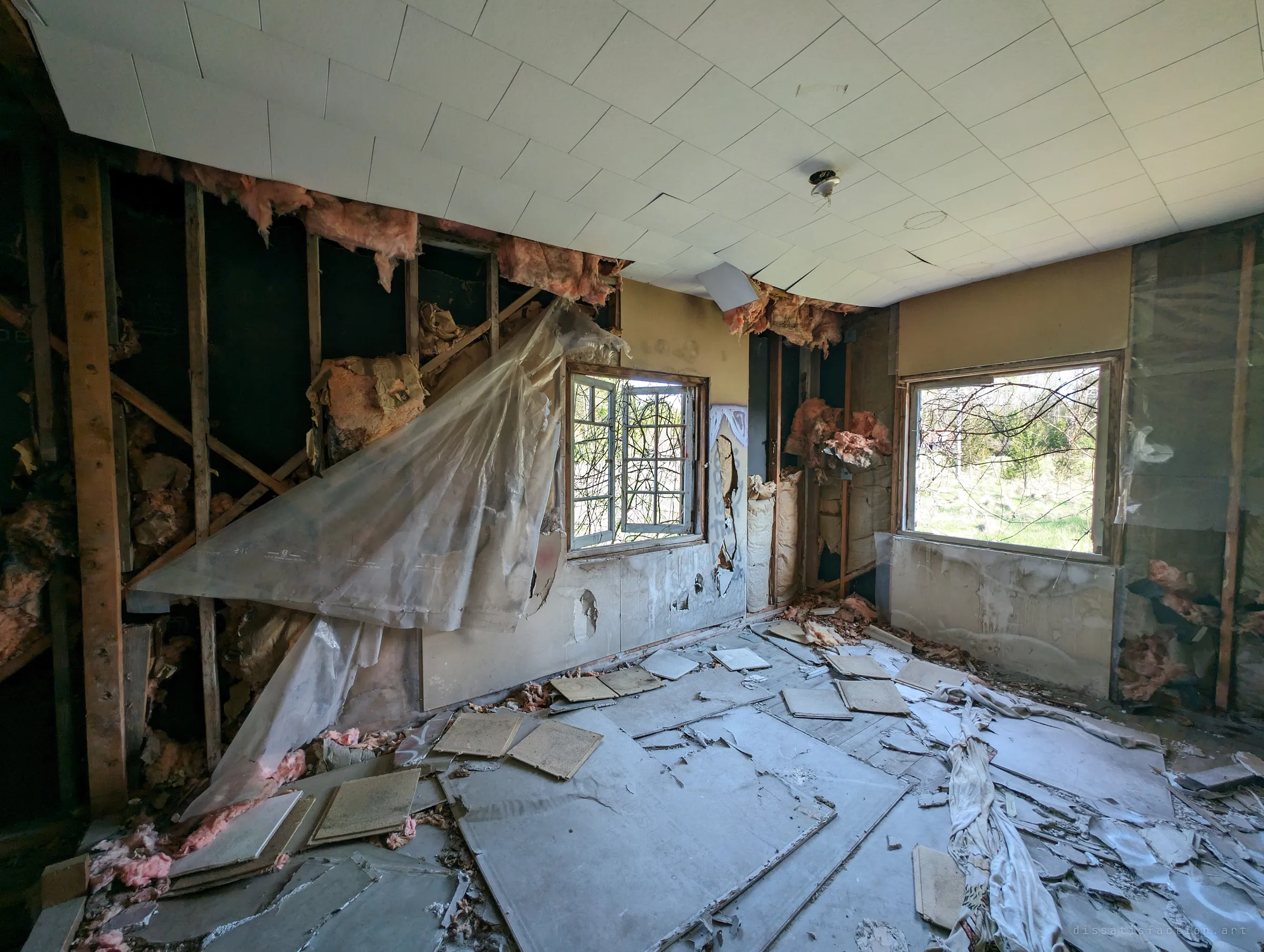 A gutted bedroom with its walls stripped to the studs, pink insulation spilling from the framing and a plastic vapour sheet hanging loose, ceiling tiles scattered across the floor. Spring light comes through two open windows onto overgrown brush.