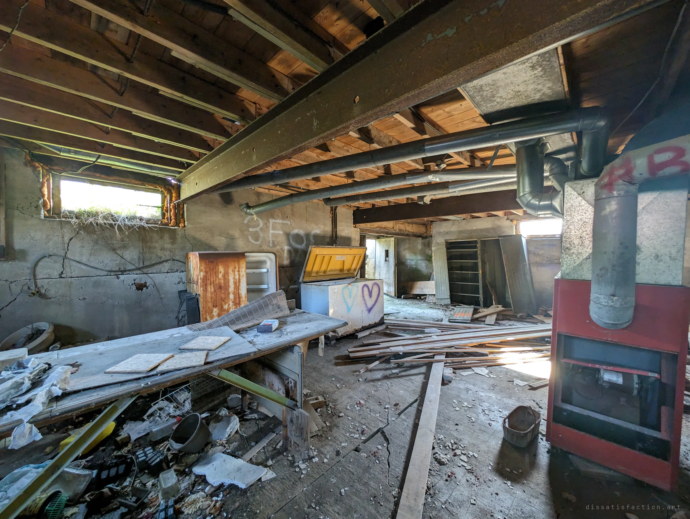 A low basement under a steel beam and ductwork, with a rusted fridge, a chest freezer sprayed with hearts and an old red furnace among scattered lumber. Daylight leaks in through a grass-level window.