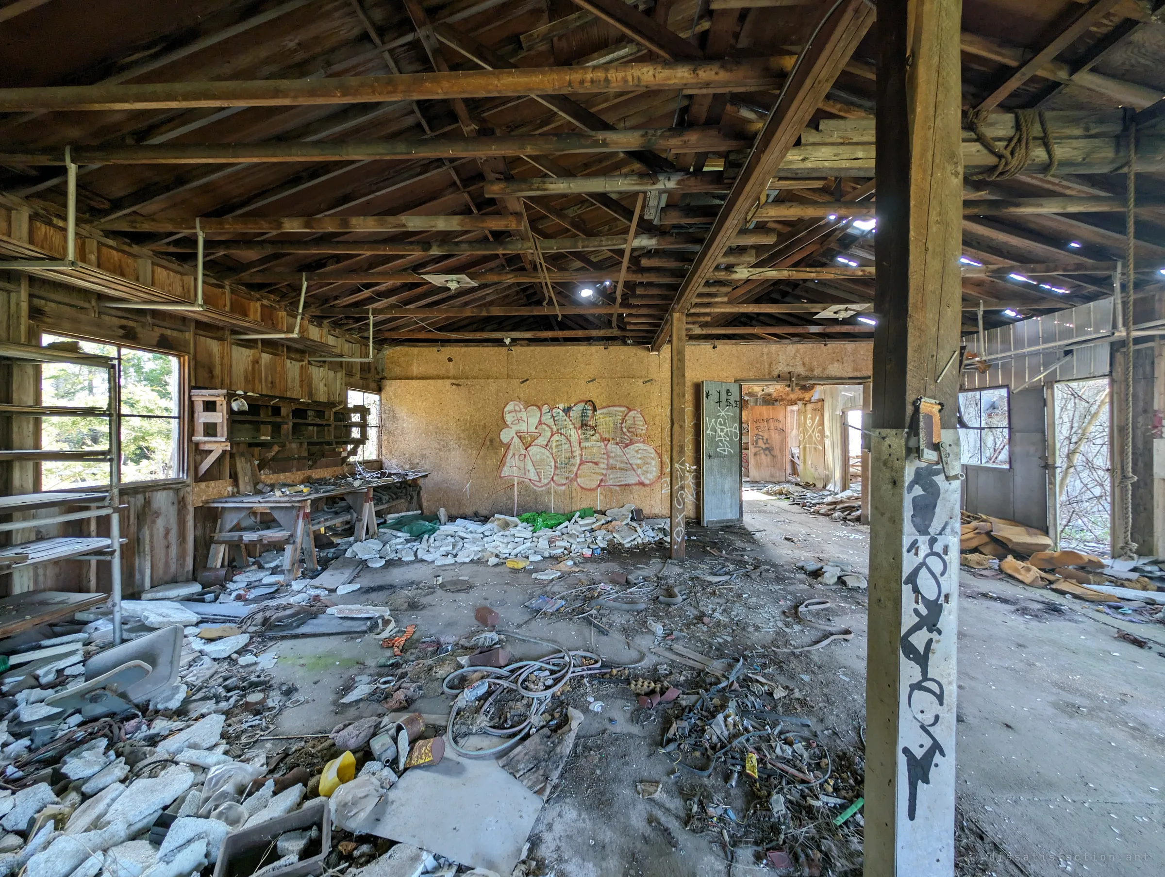 A wide timber-roofed workshop with pinholes of daylight through the roof, a graffiti piece on the back wall and floors buried in foam, wire and junk. A tagged post in the foreground leads the eye through open doors into further rooms.