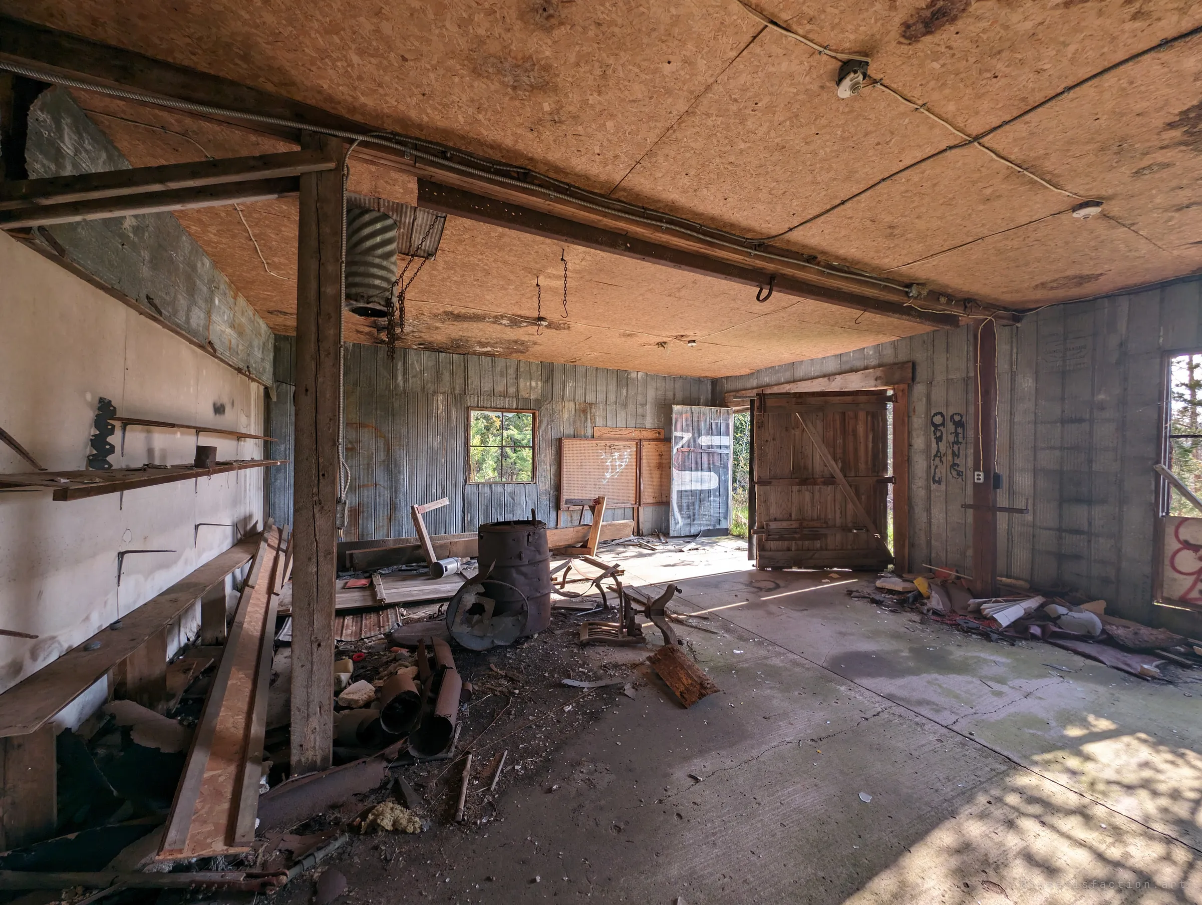 A board-walled garage with a rusted barrel stove, empty shelf brackets and pegboards, sunlight falling through an open plank door onto the concrete floor. The room feels recently abandoned rather than ruined.