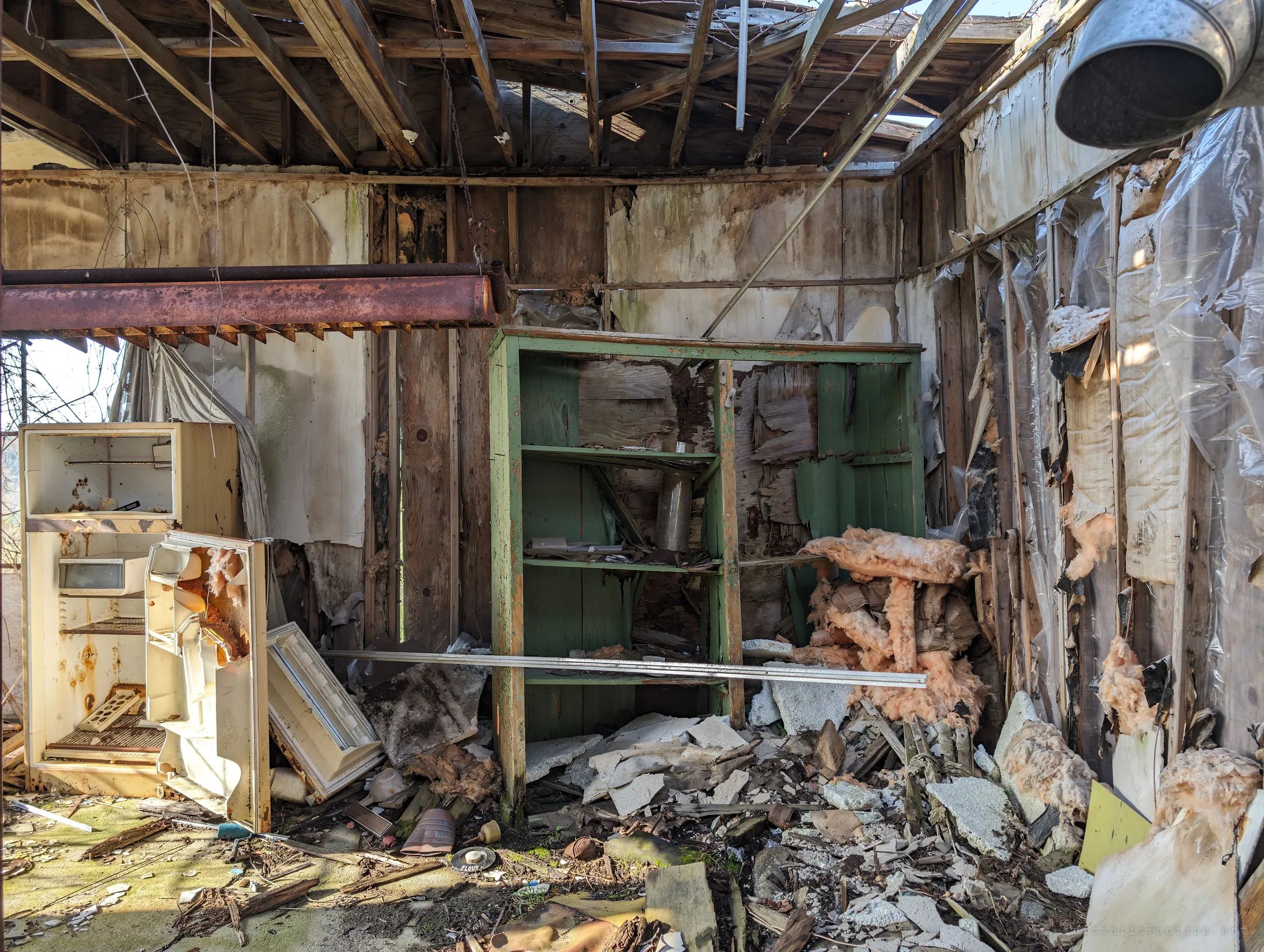 A roofless kitchen corner where an open, rust-spotted fridge stands beside a green wooden cupboard, surrounded by fallen insulation and plaster. Sun pours in through the missing ceiling.