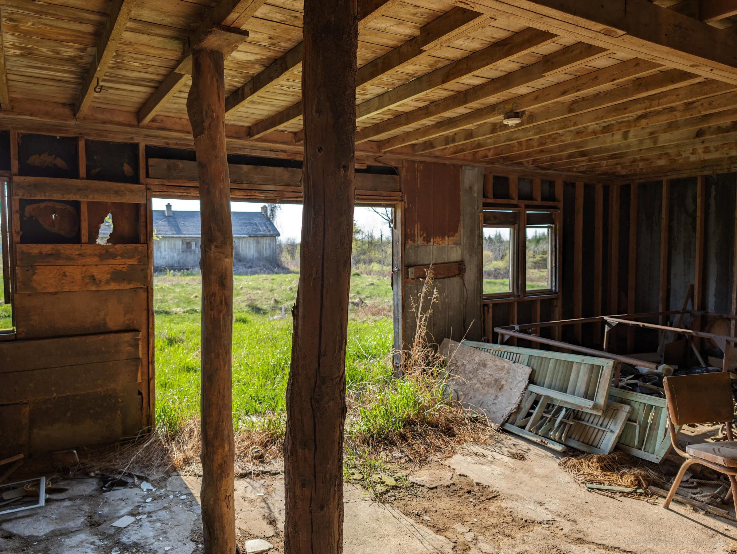 Two rough log posts hold up a plank ceiling in an open-sided shed, looking out over bright green grass to another grey farmhouse. Old shutters and a lone chair lean in the shade on the right.