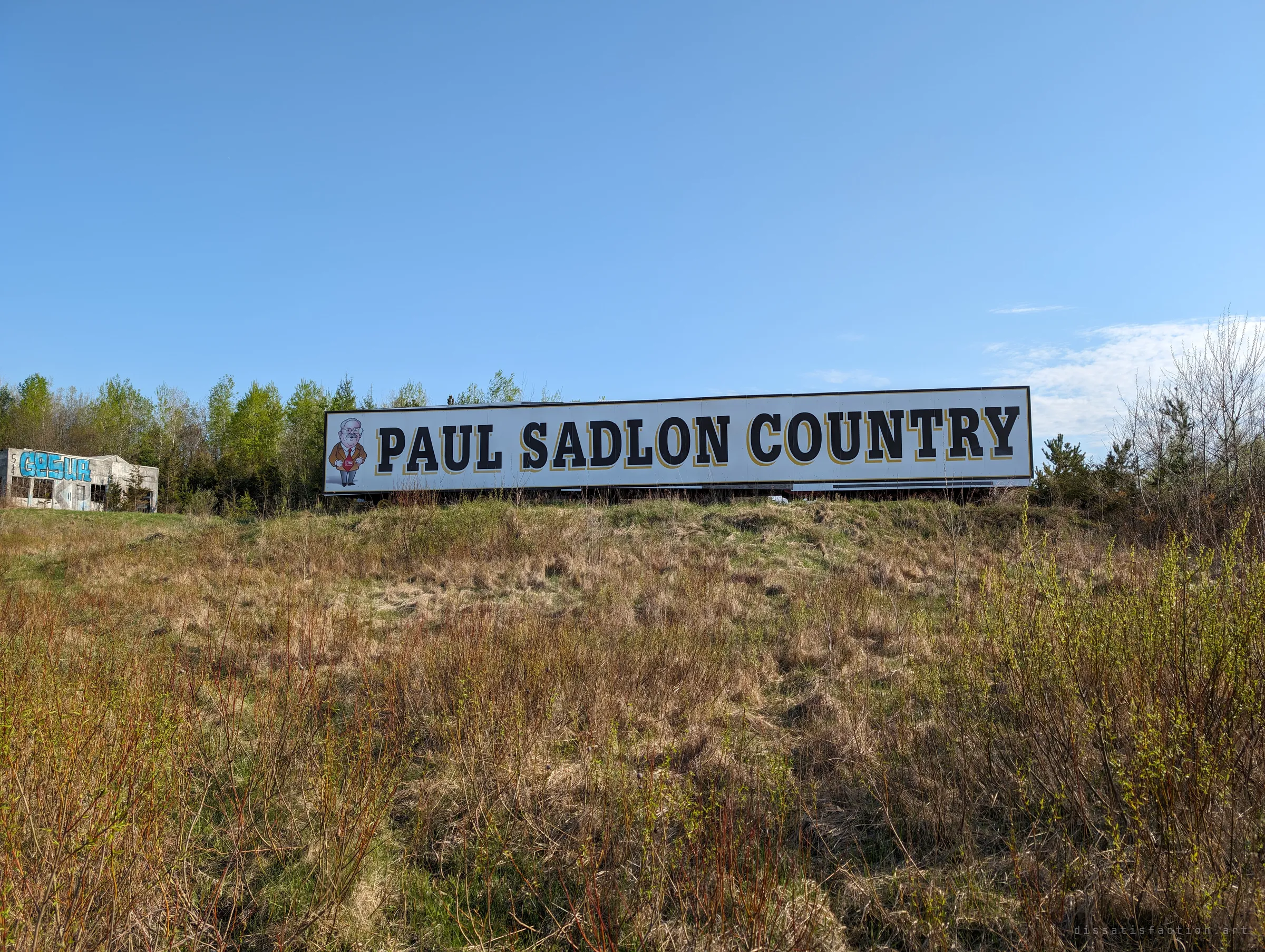 A long billboard sits on a rise above a field of dry spring grass under a clear sky, with a small graffitied ruin at the edge of the trees. It shows how the site sits beside the road, watched over by old advertising.