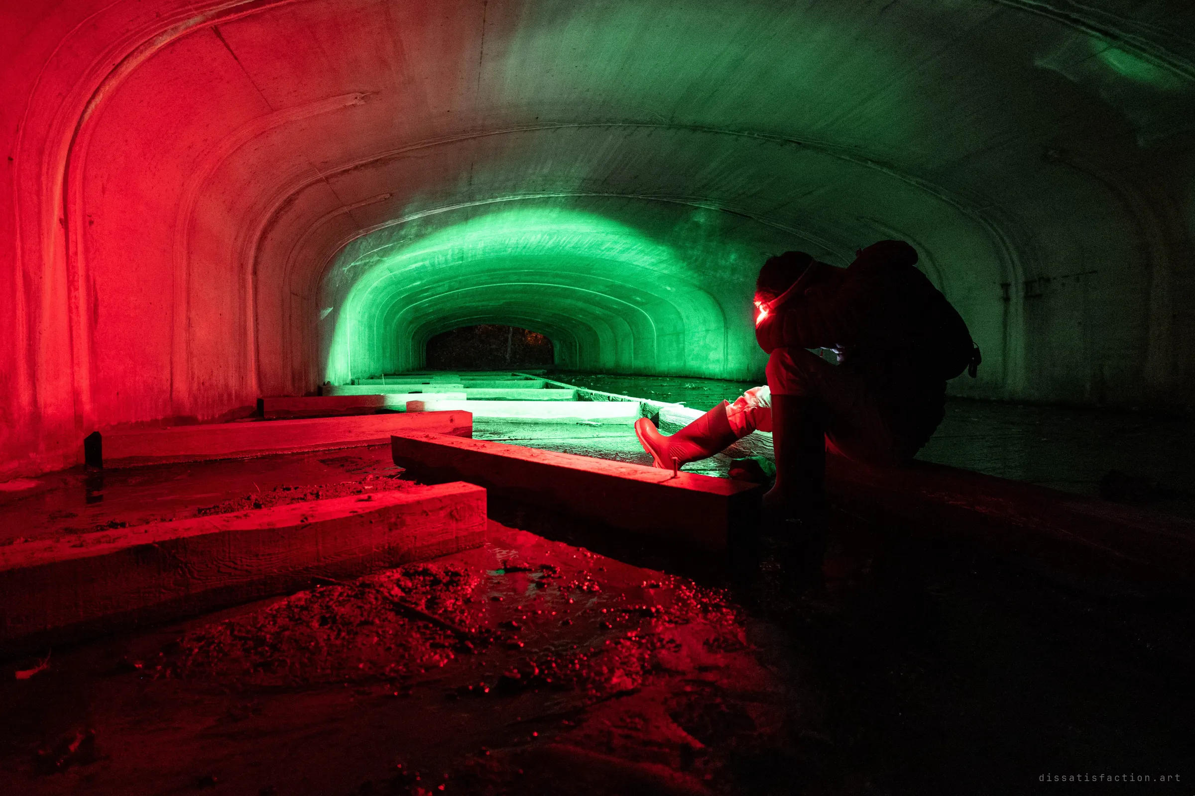 A hooded explorer in rubber boots crouches in a concrete drain tunnel with a red headlamp, the near wall washed in red light and the long arch ahead lit green.