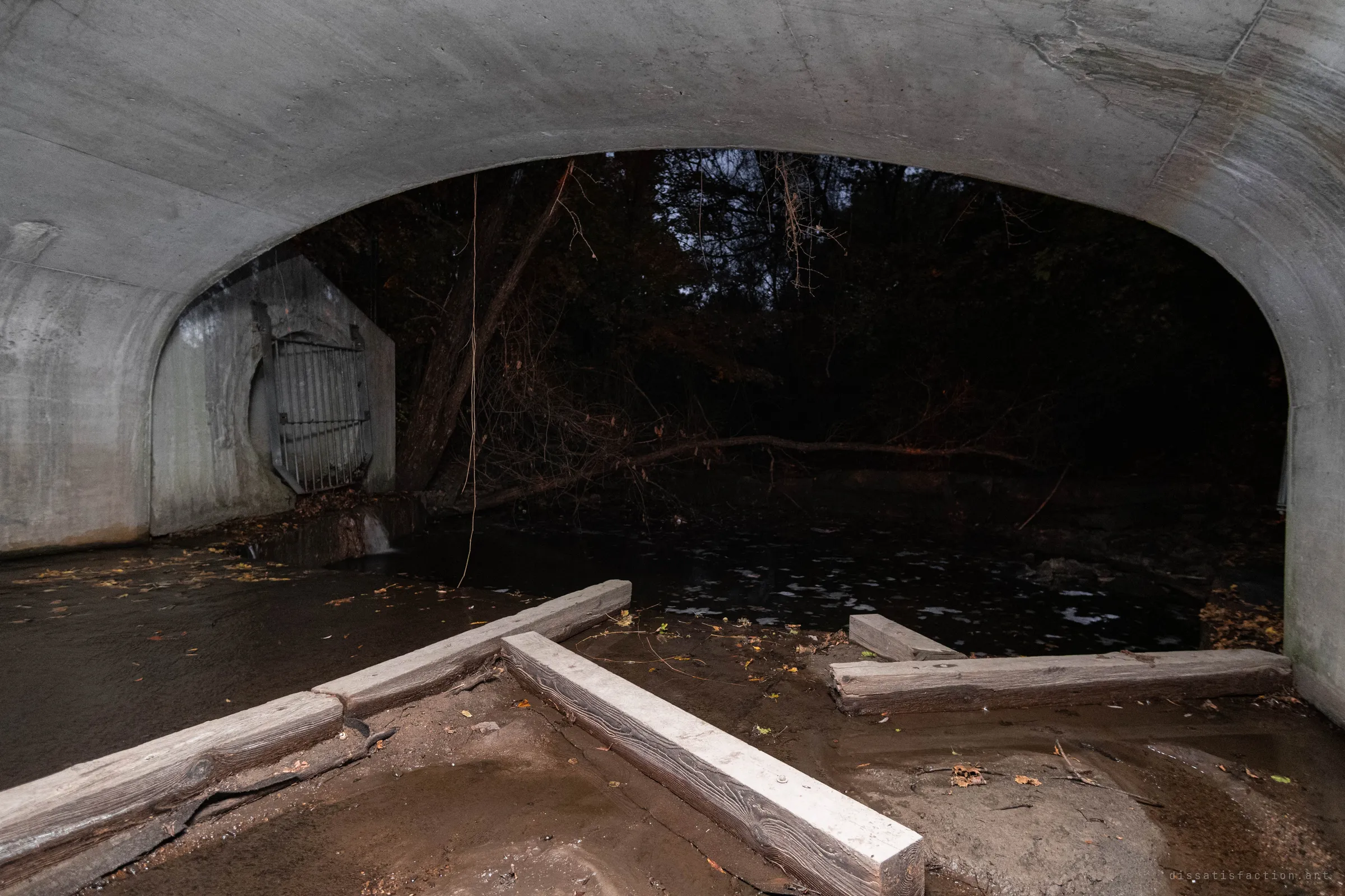 Under a wide concrete arch, a barred outfall opening sits in the wall at left above a shallow creek, with timber beams laid across the muddy bed and dark woods beyond.