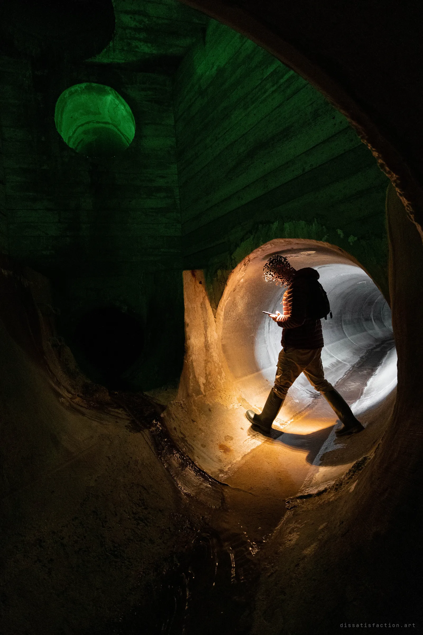 A silhouetted explorer in rubber boots walks through the lit mouth of a large concrete pipe while checking a phone, with a green-lit round opening glowing in the dark chamber behind.