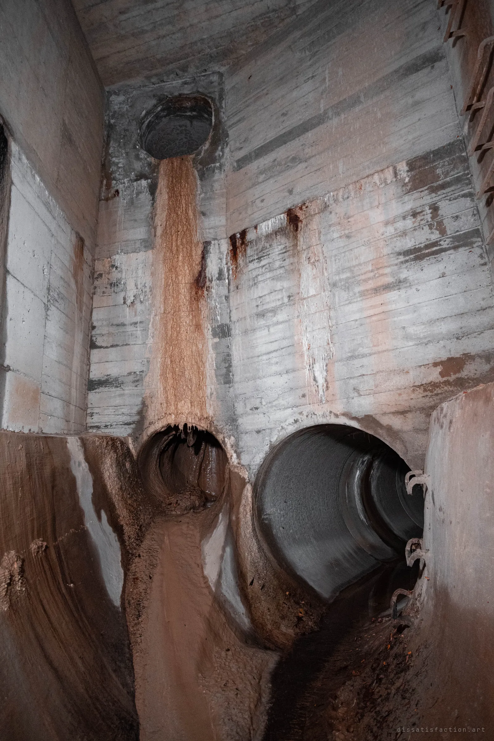 Looking up inside a concrete chamber where a rust-stained pipe pours down the wall into a muddy channel, with a second large dry pipe opening to the right.