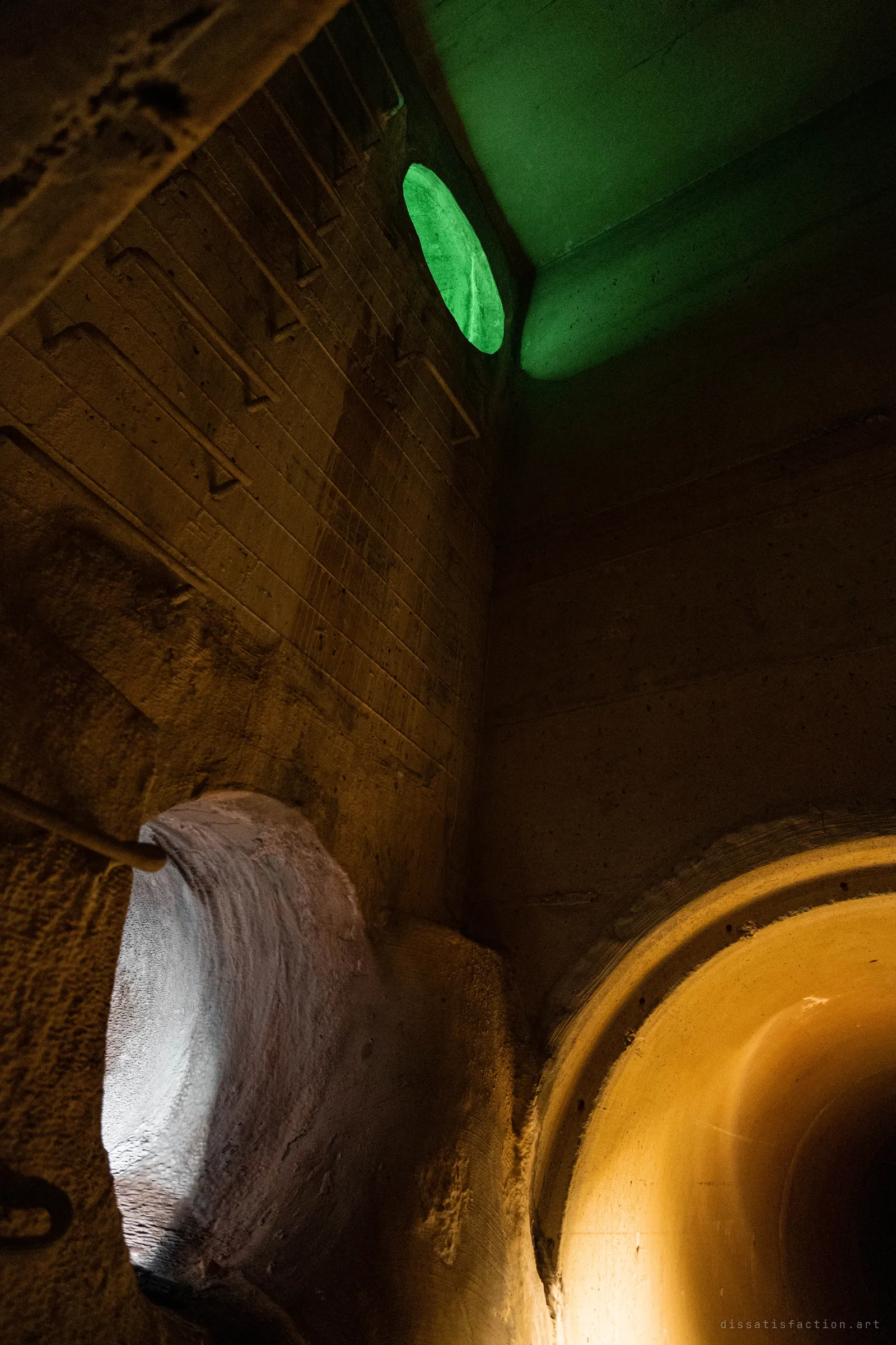 A dark concrete chamber seen from below at a tilt, with steel rungs set into the wall, a green-lit round opening above and an amber-lit pipe curving in at the lower right.