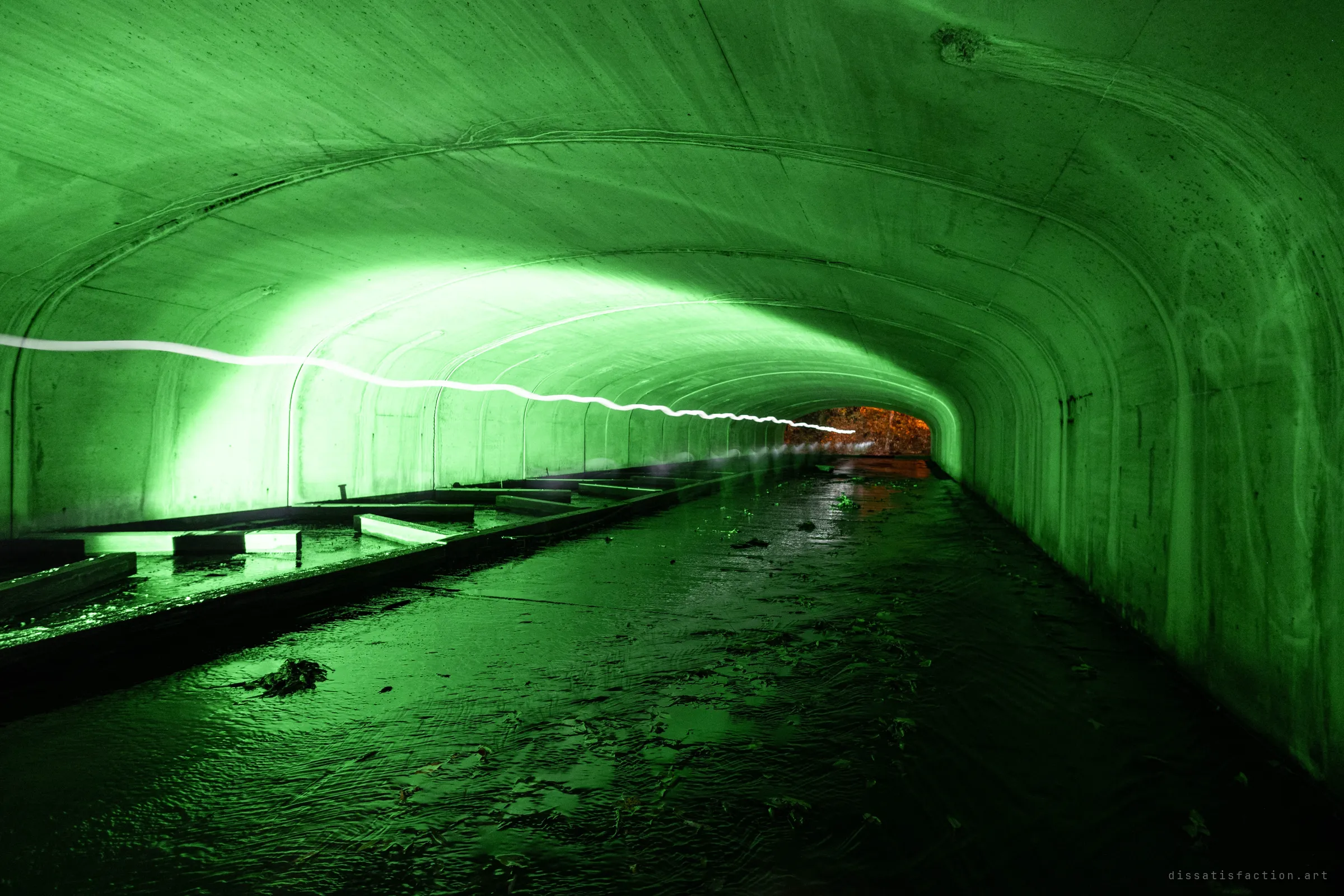 Looking down a wide arched concrete drain lit green, with a wavy white light-painting streak along the far wall and a bright autumn-coloured opening at the end.