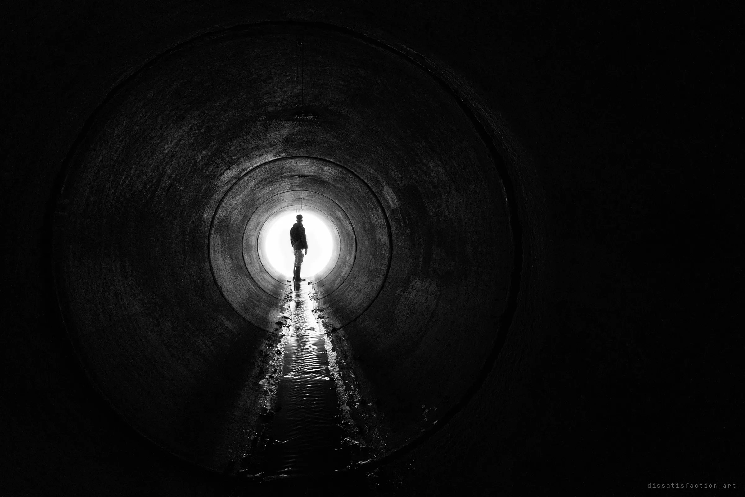 Black and white view down a huge round concrete pipe toward a bright opening, where a lone figure stands in silhouette above a thin ribbon of rippled water.